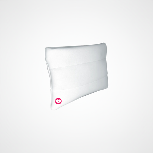A white contoured lumbar support pillow with horizontal stitched channels and a small pink lips logo near the bottom left corner, shown on a light gray background.